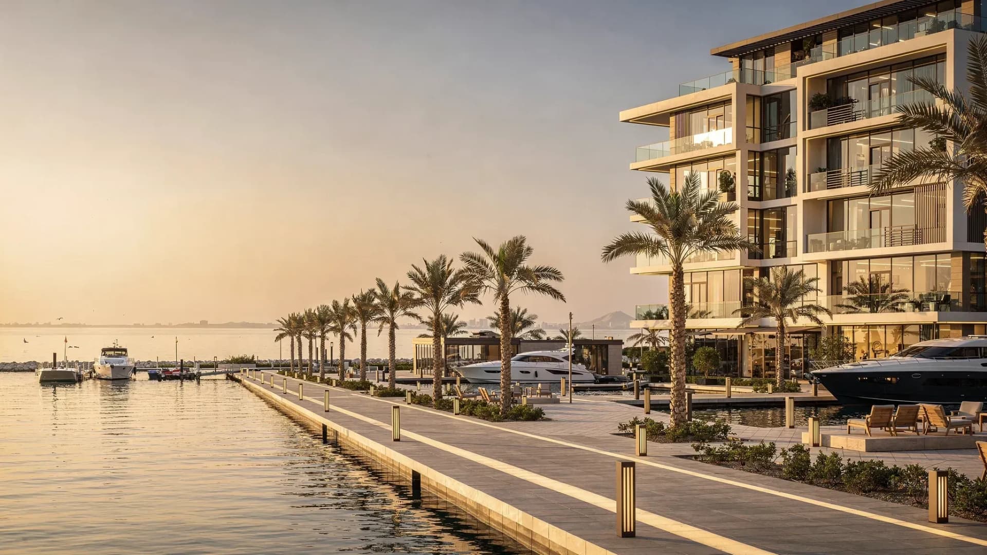 A modern luxury waterfront apartment building in Ras Al Khaimah with a calm sea view, palm-lined walkway, and nearby marina-style amenities.